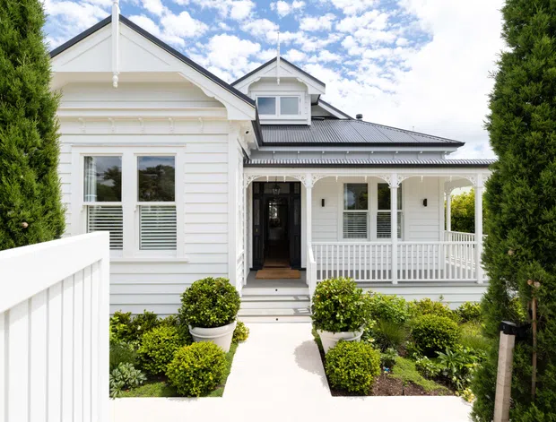 Front exterior view of a white weatherboard house with a gabled roof, dormer window, decorative verandah, white railings, and a landscaped entry path framed by tall narrow evergreen trees. The facade has a traditional heritage-inspired character with contrasting dark grey roofing and detailed timber trims.