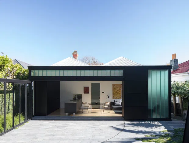 A contemporary black-clad single-storey rear extension viewed from an outdoor paved area. The extension has broad dark-framed glazed openings, translucent clerestory-style glazing, and a large sliding or folding glass door revealing a compact bright interior workspace or living area. Neighboring pitched-roof homes and lush planting are visible around the enclosed courtyard-like setting.