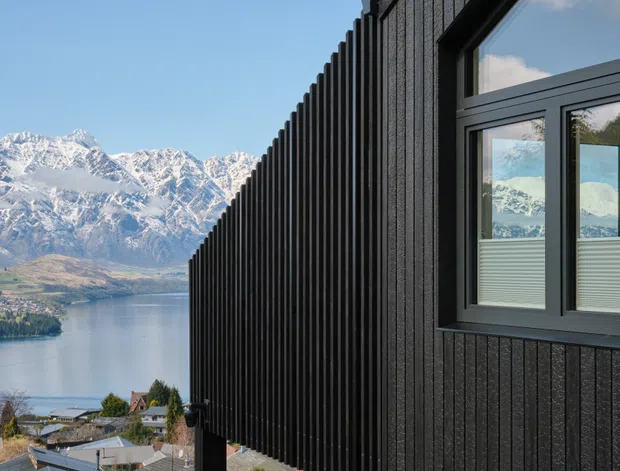 Exterior architectural view of a dark vertically ribbed building facade with black-framed glazing, overlooking a lake and dramatic snow-capped alpine mountains. The facade creates strong linear perspective, while reflections in the windows echo the mountain setting.