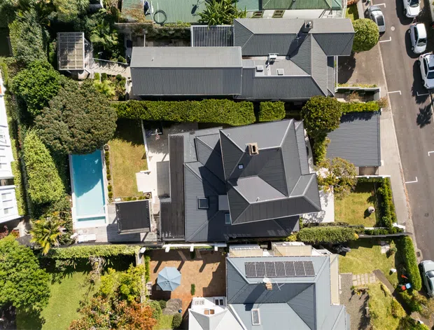 Aerial top-down view of a dense suburban residential neighbourhood, showing several detached homes with dark pitched metal roofs, landscaped gardens, hedges, paved outdoor areas, and a rectangular swimming pool. Solar panels are visible on one roof, with a street and parked cars along the right edge.