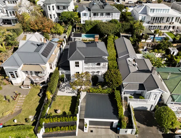 Aerial view of a dense residential neighbourhood featuring closely spaced detached homes, predominantly two-storey houses with pitched and gabled roofs, light-coloured façades, landscaped gardens, decks, driveways, and several visible swimming pools. The homes display a mix of contemporary and traditional suburban architectural forms, including dark metal roofing and white-painted exterior detailing.
