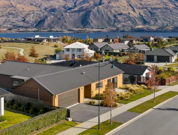 Aerial view of a modern suburban residential neighbourhood with detached homes, broad paved streets, landscaped verges, and a lake or body of water backed by rugged mountains. The foreground includes a single-storey home with dark pitched metal roofing, warm brick walls, and an attached garage, surrounded by lawns and young trees.