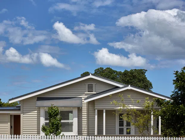 Front exterior view of a single-storey weatherboard house with a prominent gabled roof, covered front porch, white picket fence, and neatly maintained suburban frontage. The facade is painted a light neutral grey-beige with white trim and a dark grey roof. Blue sky and trees form the background.