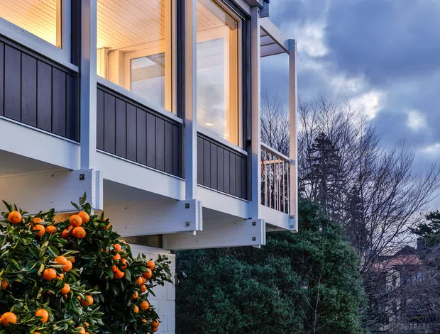 Exterior architectural photograph of a modern elevated residential building at dusk. The facade features large floor-to-ceiling glazed openings with dark vertical cladding or panels, white structural framing, and a projecting balcony or covered outdoor platform with a simple metal railing. Warm interior lighting contrasts with the cool blue-grey sky. Citrus trees and landscaped paths surround the building, with neighbouring homes and mature vegetation in the background.