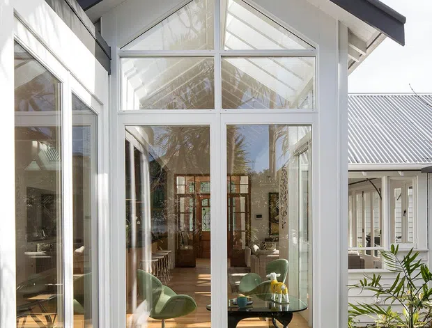 Exterior view of a bright white weatherboard house extension or sunroom with a steep gabled roof, large floor-to-ceiling glazed wall, and white-framed windows and doors. The glazing reveals a furnished interior with green lounge chairs and timber flooring. A small planted courtyard with concrete stepping stones sits in the foreground, while corrugated metal roofing is visible on the adjoining structure.