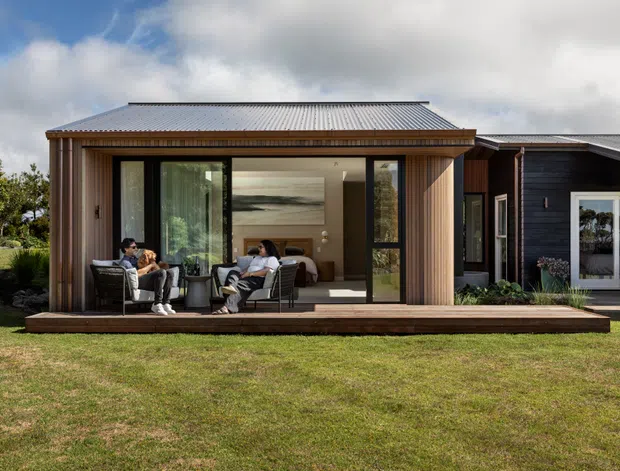 Single-storey contemporary modular-style dwelling with a corrugated metal gable roof, vertical timber cladding, and large glazed openings. The open front reveals a compact living area with two lounge chairs, a small table, built-in cabinetry and a bed-like sleeping area, while a darker timber-clad adjoining volume is visible to the right. The building sits on a simple timber deck within a broad grassy landscape.