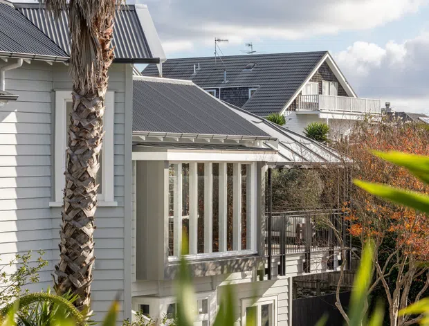 Exterior view of a light-painted, multi-level residential building with overlapping pitched metal roofs, white-framed glazing, balconies, and weatherboard-style cladding. A palm tree and landscaped planting partially frame the foreground, while neighbouring houses are visible behind.