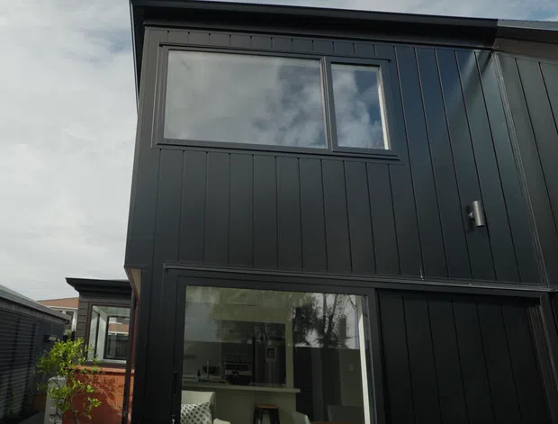 Exterior view of a modern two-storey building clad in dark vertical panels, with large rectangular windows reflecting the sky and surrounding trees. A glazed lower-level opening reveals part of an interior living space, while neighbouring structures and a small planted area are visible at the edges.
