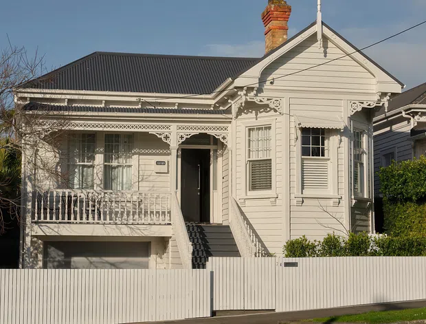 Street-facing view of a restored single-storey Victorian-era timber cottage with ornate lacework verandah, decorative gable detailing, weatherboard cladding, corrugated metal roofing, brick chimney, and a raised entry above an undercroft garage or storage area. The house is painted a pale grey-white and enclosed by a simple corrugated metal front fence.