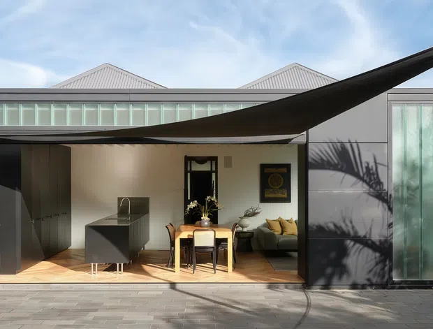A contemporary single-storey house viewed from an outdoor paved area, with a broad open glazed frontage revealing a compact kitchen, dining area and living space. The building has corrugated metal gabled roof forms, dark exterior cladding, large glass doors and windows, and a prominent black triangular shade sail across the facade.