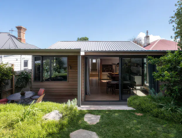Exterior view of a compact single-storey residential extension or garden studio with horizontal timber cladding, dark-framed full-height glazing and a corrugated metal roof. A covered outdoor patio with a small table and chairs sits in front, opening onto a landscaped garden. The glazed doors reveal a warm interior with built-in cabinetry and dining furniture. Neighbouring rooflines and chimneys are visible behind the structure.