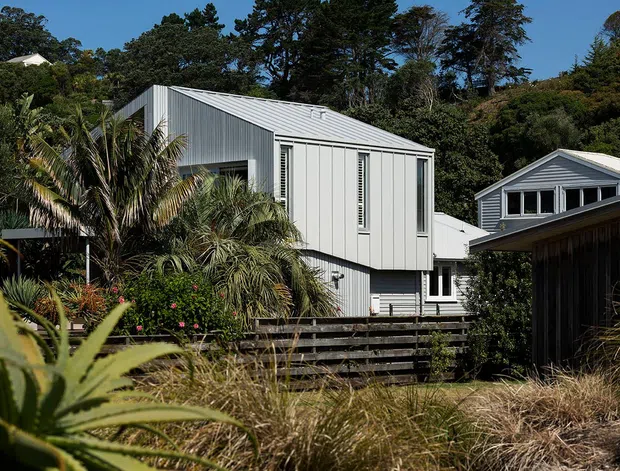 Exterior view of a contemporary raised home in a leafy suburban or coastal setting. The building has pale grey vertical corrugated cladding, a matching pitched metal roof, large glazed openings with louvred or screened windows, and timber decking and retaining fences. Tropical and native planting surrounds the house, with neighbouring pitched-roof buildings visible in the background.