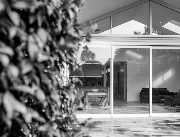 Black-and-white exterior photograph of a contemporary house viewed from the garden. A large glazed living area opens onto a paved terrace, with a pitched roof, high clerestory glazing, and a visible modern sofa and dining area inside. Dense foreground planting partially frames the facade, while lawn occupies the foreground.