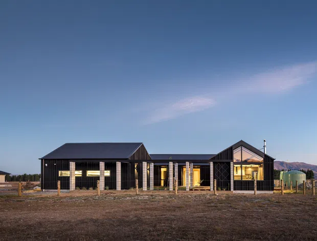 A low, single-storey contemporary rural residence photographed from the front at dusk. The long dark-clad building has multiple gabled roof volumes, extensive glazing, timber posts and a central recessed entry or connection. Open bare ground and fencing surround the house, with distant mountains visible behind it.