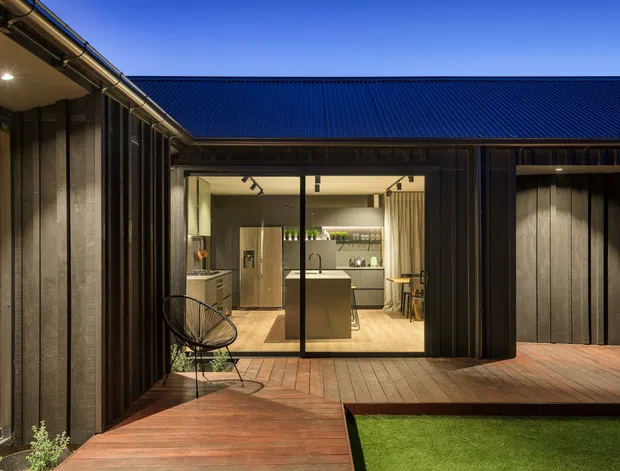 Twilight exterior view of a dark-clad contemporary home arranged around a courtyard or deck. The image looks toward a brightly lit kitchen through large glazed sliding doors, with a central island, pale cabinetry and timber flooring visible inside. Timber decking, a small outdoor chair, a strip of lawn, vertical dark cladding and a corrugated pitched roof define the exterior.