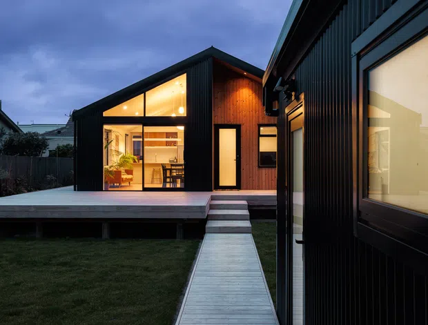 Exterior dusk view of a compact contemporary house with dark vertical cladding, warm timber accents, and expansive illuminated glazing. A raised timber deck and narrow timber walkway lead through a lawn to the entrance, while the interior kitchen and dining area are visible through the glass.