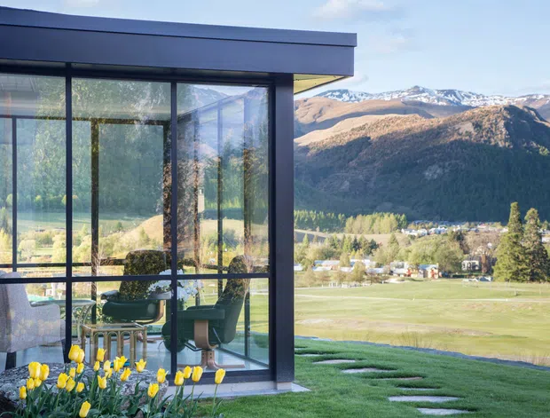 Modern single-storey glazed building extension or pavilion with dark metal framing and a flat roof, opening onto a manicured lawn. The transparent corner glazing reveals a compact seating area inside, while a broad mountain landscape with snow-capped peaks is visible beyond.