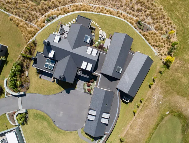 Aerial view of a large, dark-roofed multi-wing residential house set within expansive landscaped grounds. The building has several connected pitched roof forms, skylights, roof windows, paved driveways, curved garden paths, and surrounding ornamental grasses and lawns.