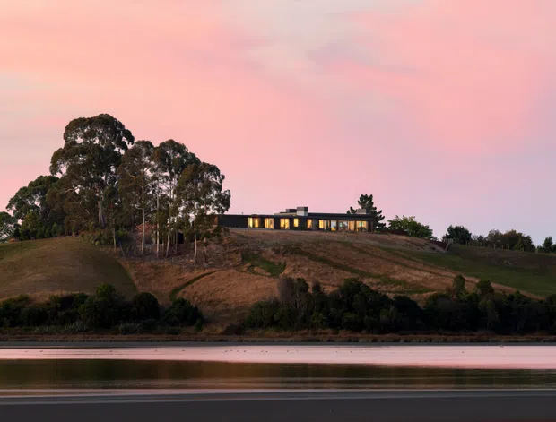 A low, modern single-storey house sits along the crest of a gently sloping rural or semi-rural hillside, viewed across calm water at dusk. The dark horizontal building has a flat roof and a continuous band of warm illuminated glazing, partially screened by mature trees and native-looking planting. The scene has a tranquil, minimal composition with pink evening sky and reflected water in the foreground.