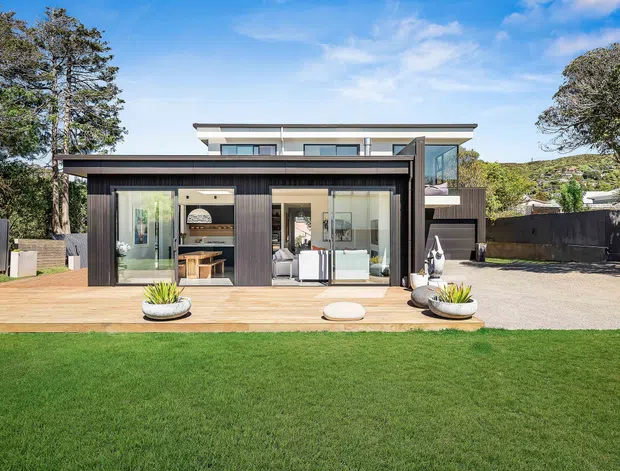 A contemporary two-storey residence viewed from the rear garden, with dark vertical cladding, broad floor-to-ceiling glazing, and a timber deck opening to an open-plan interior. The upper level has a light-toned flat roof and glazed corner, while the property includes paved outdoor space, landscaped lawn, mature trees, and sculptural planters.
