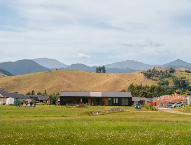 Wide exterior view of a low, modern single-storey rural property set in an expansive green field, with several nearby buildings and rolling golden hills and mountains in the background. The main residence has a long horizontal form, dark cladding, a low-pitched roof, and generous glazing. The scene has an open agricultural or rural setting and a restrained contemporary appearance.