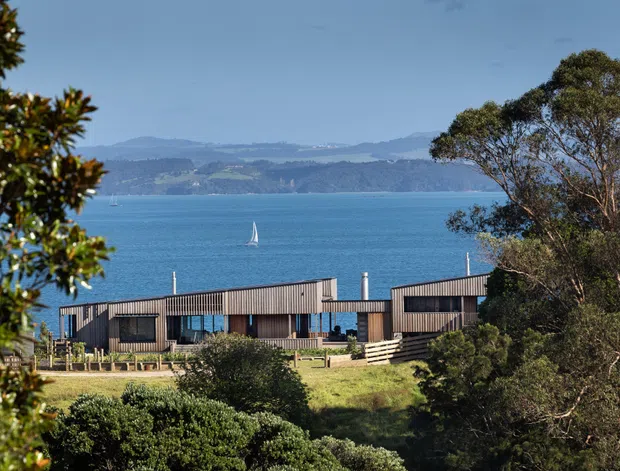 Exterior view of a low-rise modern building set on a landscaped site overlooking blue water, with a distant mountainous coastline and sailboat beyond. The building has a long, angular form with dark vertical cladding, extensive glazing, and multiple pitched roof sections, partially screened by mature trees and shrubs.