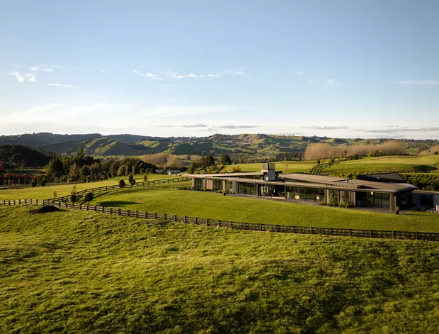Wide exterior landscape view of a low, elongated contemporary building set within expansive green rural farmland. The property has a largely flat roofline, extensive glazing, dark structural elements, and fenced paddocks, with rolling hills and cultivated fields in the background under a clear sky.