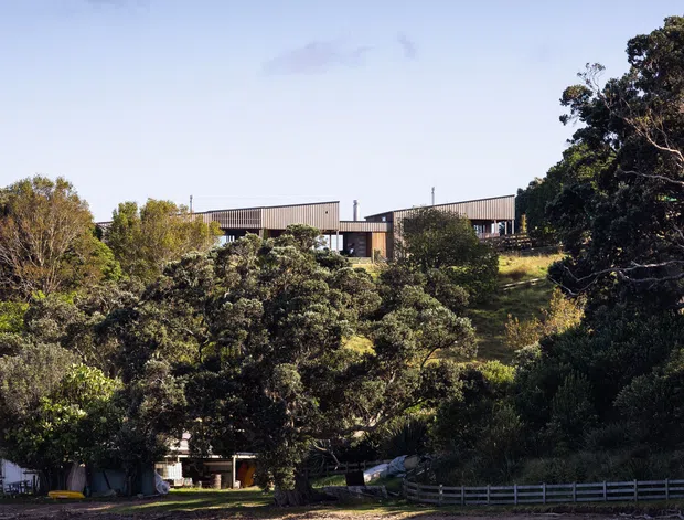 A distant exterior view of a low-profile modern house set on a wooded, gently sloping waterfront site. The building has long horizontal forms, dark vertical cladding, broad roof planes, and large areas of glazing, partially screened by dense trees. A pebbly shoreline and calm blue-green water occupy the foreground.