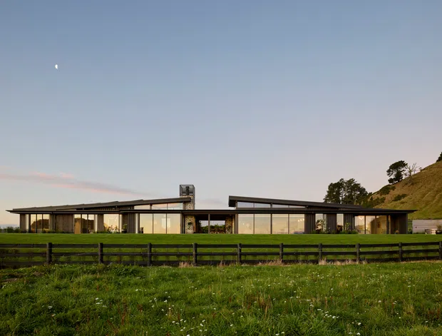 A low-slung contemporary single-storey house set in a rural, gently sloping landscape. The building has a broad flat or very low-pitched roof, extensive floor-to-ceiling glazing, dark slender structural framing, and warm timber-toned wall elements. A dark post-and-rail fence and grassy foreground frame the view, with a small moon visible in the open sky.
