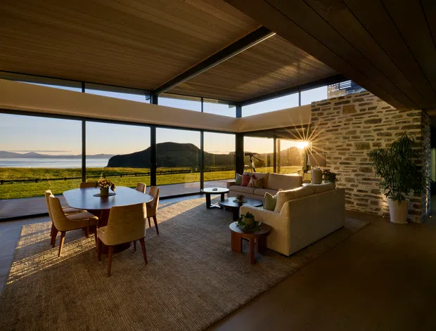 A warm, open-plan living and dining space with expansive floor-to-ceiling glazing overlooking a coastal landscape and water. The room features a timber-lined ceiling with recessed linear lighting, dark slender window frames, a natural stone feature wall, timber flooring, a neutral sectional sofa, and a round dining table with upholstered chairs. The low evening sun creates a relaxed, inviting atmosphere.