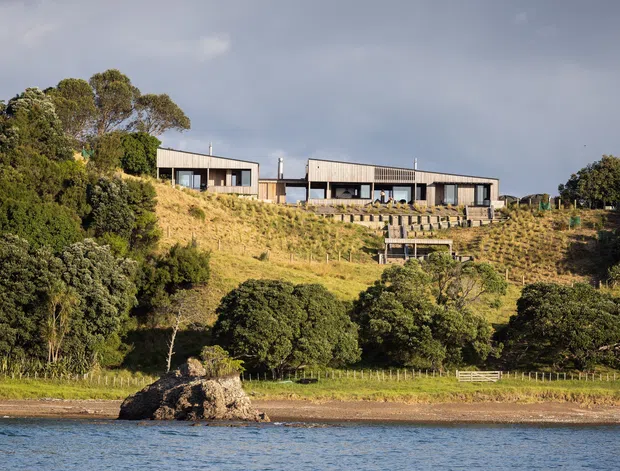 Wide exterior view of a long, low-slung contemporary house set on a grassy, tree-covered hillside above a body of water. The residence has strong horizontal lines, dark framed glazing, pale timber or weatherboard-like cladding, and a broad terrace or deck. The image is photographed from across the water, with a rocky shoreline and mature vegetation in the foreground under a cloudy sky.