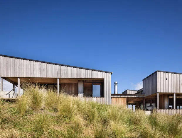 Exterior view of a contemporary low-rise coastal home composed of interconnected rectangular volumes. The building features vertical weathered timber cladding, dark metal roof edges and framing, covered verandah-like outdoor spaces, and large glazed openings. Native dune grasses occupy the foreground beneath a clear blue sky.