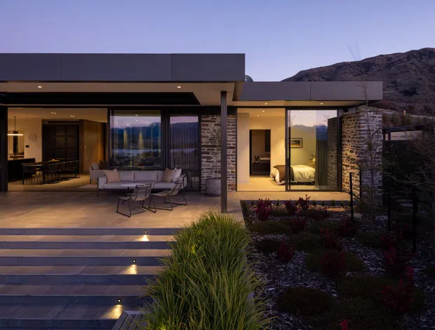 Twilight exterior view of a contemporary single-storey home set against a rugged mountain backdrop. The flat-roofed structure has extensive floor-to-ceiling glazing, dark metal or rendered wall planes, stone-clad columns and walls, and a broad stepped terrace. Through the open living area, a dining zone and lounge are visible, while a bedroom is visible through glazing on the right. The landscaped foreground includes ornamental planting and integrated step lighting.