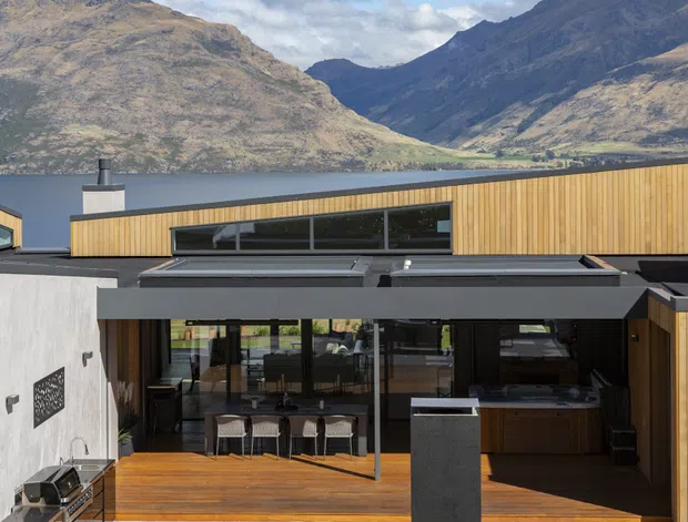 A contemporary lakeside residence viewed from the outdoor terrace, with a low-profile timber and dark metal facade, extensive glazing, and an open-plan kitchen and dining area inside. The home overlooks a dramatic mountainous lake landscape. A built-in outdoor grill and paved entertaining area are visible in the foreground.