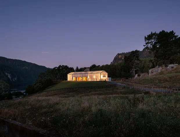 A modern illuminated house set into a rugged, elevated rural landscape at dusk. The low-profile building has extensive warm-lit glazing, a simple dark roofline, and appears surrounded by natural grasses, rocky terrain, forested hills, and mountain cliffs.