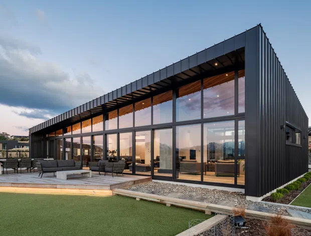 Exterior view of a contemporary single-storey building with a long glazed facade, dark vertically ribbed cladding, and a projecting flat roof. The interior is visible through floor-to-ceiling glazing, while a paved outdoor terrace with lounge furniture fronts a landscaped lawn.
