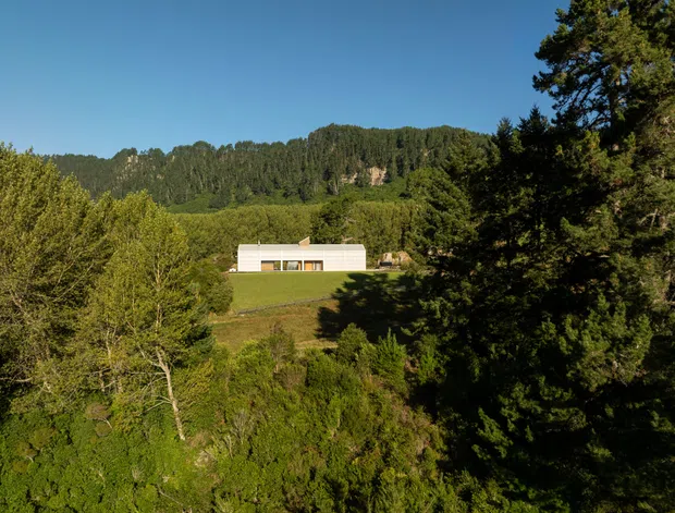 A distant exterior view of a minimalist rectangular house set on a grassy clearing amid dense forest, with a wooded mountain ridge behind it. The low white building has a simple flat or very low-pitched roof, horizontal glazing, and a strong connection to the secluded natural landscape. Bright daylight and extensive greenery dominate the scene.