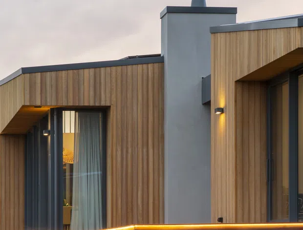 Close-up exterior view of a contemporary building with warm vertical timber cladding, dark metal-framed glazing, projecting roof edges, a rendered chimney volume, and integrated warm wall lighting. A timber-clad terrace or privacy wall with linear lighting runs across the foreground.