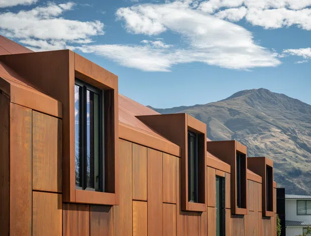 Exterior architectural photograph of a contemporary low-profile building with repeating angular volumes, narrow vertical windows, and a weathered rusty-orange metal-clad facade and roof. A dramatic mountain rises behind the building under a partly cloudy blue sky.