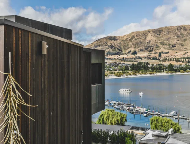 Exterior view of a contemporary dark-clad building overlooking a lake and marina, with rugged mountains and low-rise development across the water. The facade features vertical dark timber-like cladding, a flat roof, minimal openings, and a projecting upper volume.