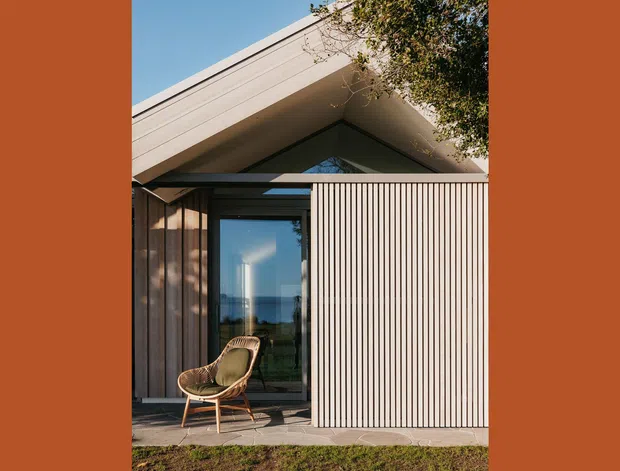 Exterior view of a contemporary single-storey home with a prominent pitched roof and triangular glazed gable. Vertical timber-look screening and dark-framed glazing form the facade, with a small paved terrace and outdoor chair in front. The image is framed by warm terracotta side borders and partial landscaping.