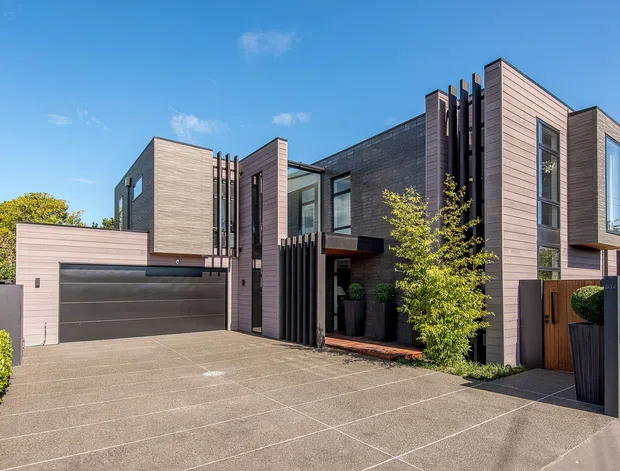 Contemporary multi-level residential facade with a broad concrete-paved driveway, integrated double garage, projecting box forms, vertical dark slat screens, and extensive horizontal timber-look cladding in muted grey and mauve tones. Large windows and a recessed entry are framed by minimalist landscaping and boundary fencing.
