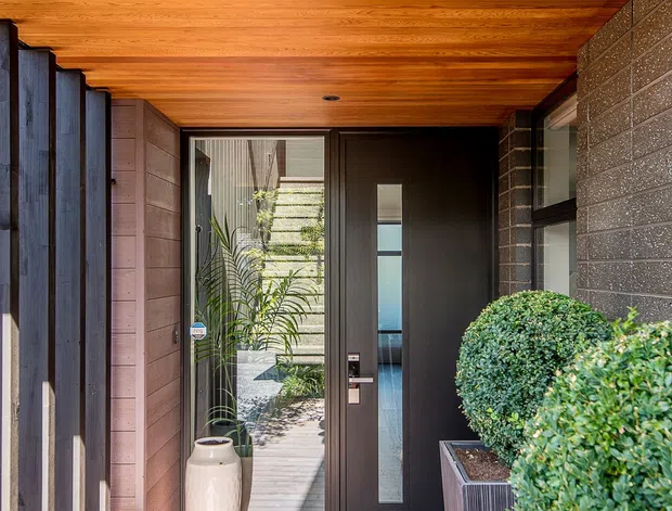 A covered residential entryway with a dark contemporary front door featuring a narrow vertical glazed panel. Warm timber boards line the ceiling and floor, while dark brick and timber-clad walls frame the entrance. Tall glazed openings, a sculptural ceramic planter, and neatly clipped greenery in raised planters create a refined, welcoming approach.