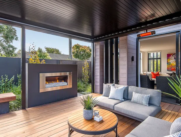 A covered outdoor lounge and entertaining area with timber decking, a built-in linear gas fireplace set into a dark feature wall, a grey outdoor sofa and coffee table, and large sliding glass doors opening into the interior. The space is framed by dark metal elements and a slatted ceiling, with planted garden screening around the perimeter.