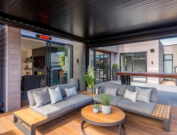 A covered outdoor lounge terrace with two grey cushioned sofas, a timber coffee table, and built-in timber decking. The space is enclosed by dark-framed glazing and a dark slatted ceiling, with a contemporary pale-clad building visible around the courtyard. Potted plants and a raised timber bar counter add function and warmth.