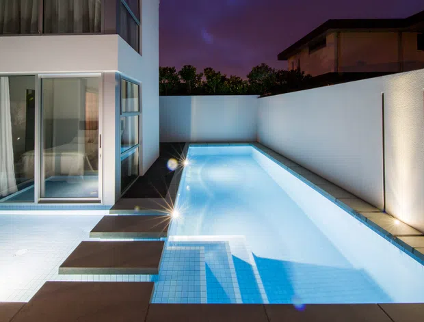 Dusk view of a modern residential pool courtyard, with a long illuminated rectangular swimming pool, tiled steps, dark paving, white rendered boundary walls, and a contemporary glass-fronted multi-storey house. Architectural lighting creates a cool blue and purple atmosphere.