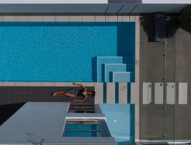 Aerial view of a contemporary residential pool area with a rectangular turquoise swimming pool, integrated shallow step section, dark timber decking, pale tiled paving, and a compact outdoor dining area beside the house.
