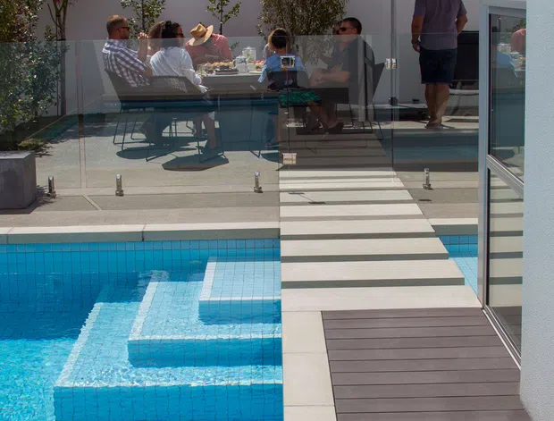 A contemporary outdoor pool area beside a modern white house, featuring vivid blue tiled water, stepped pool sections, dark decking, pale paving, frameless glass balustrades, and a raised entertaining terrace with a dining table and large dark umbrella. Several people are gathered outdoors, with landscaped planting and neighbouring buildings visible in the background.