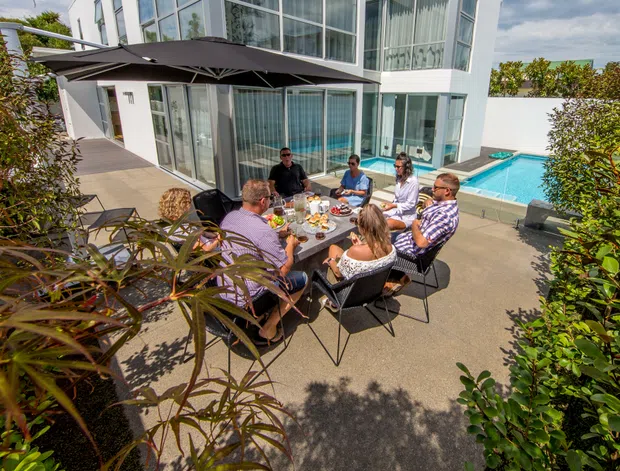 A contemporary multi-level home with extensive floor-to-ceiling glazing, a paved outdoor dining terrace, and a bright rectangular swimming pool. Several people are gathered around an outdoor dining table beneath a large cantilevered umbrella, with landscaped planting framing the foreground.