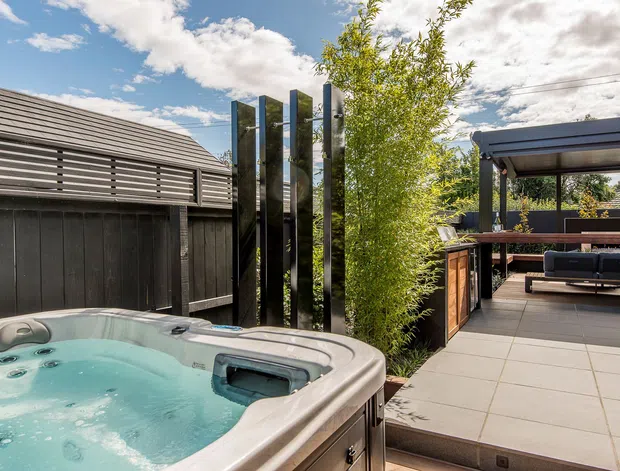 A landscaped residential outdoor entertaining area featuring a bubbling spa pool in the foreground, large-format tiled paving, a covered timber outdoor lounge and dining structure, dark vertical screening panels, and a slatted dark-clad structure. The space has a contemporary, privacy-focused design with greenery and a bright open sky.